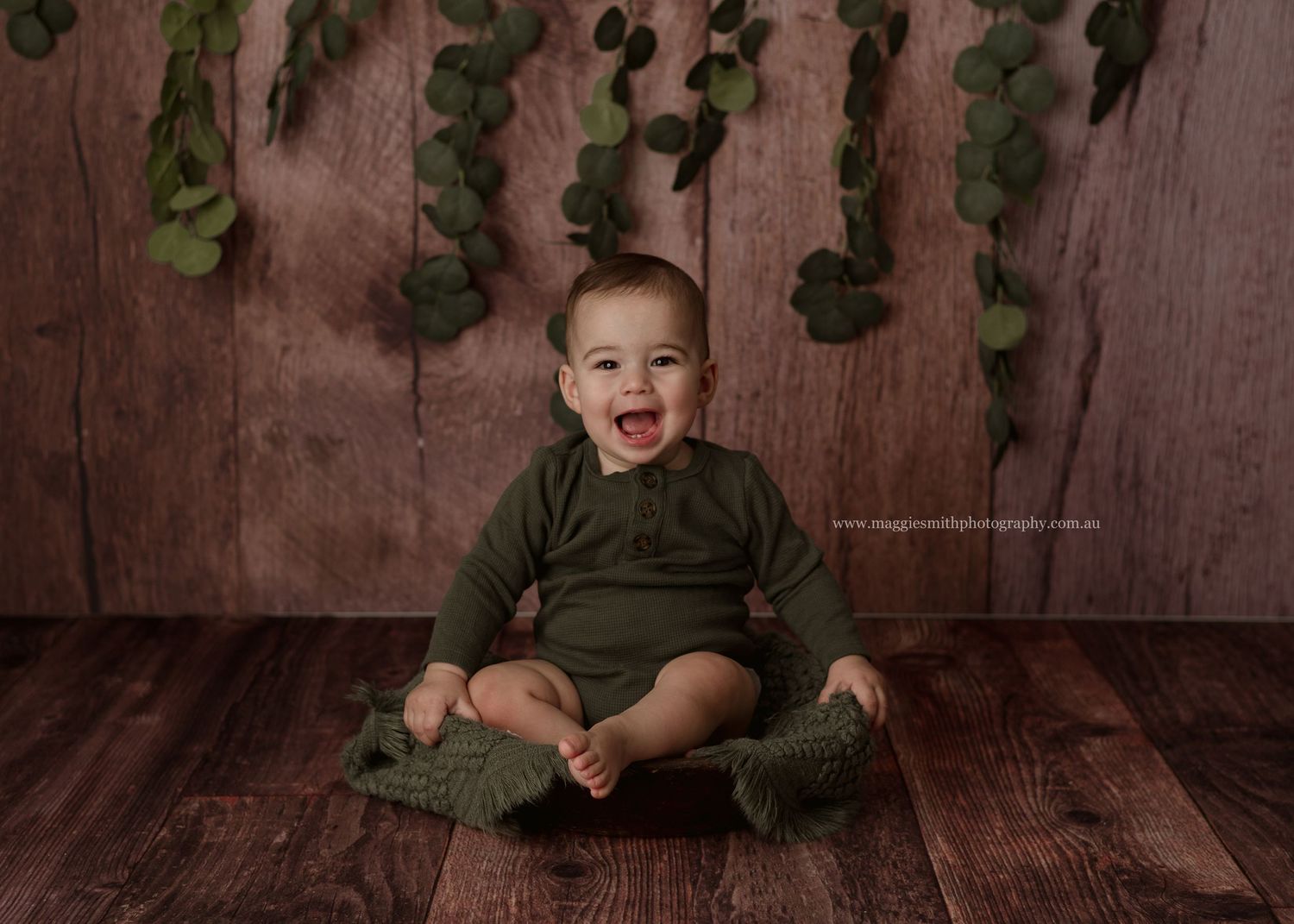A small child in olive green clothing sits on wooden floor with green hanging decorations in the background.