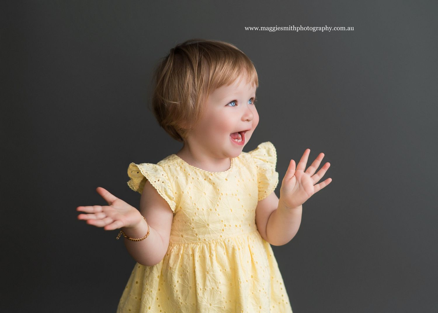 Toddler in yellow dress laughs and dances joyfully against dark grey studio backdrop. Kids photography