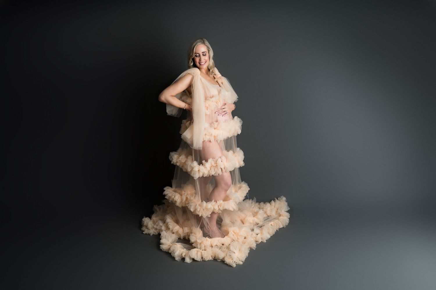 A pregnant woman poses in a layered, ruffled champagne-colored gown against a dark studio backdrop. Maternity photography fine art