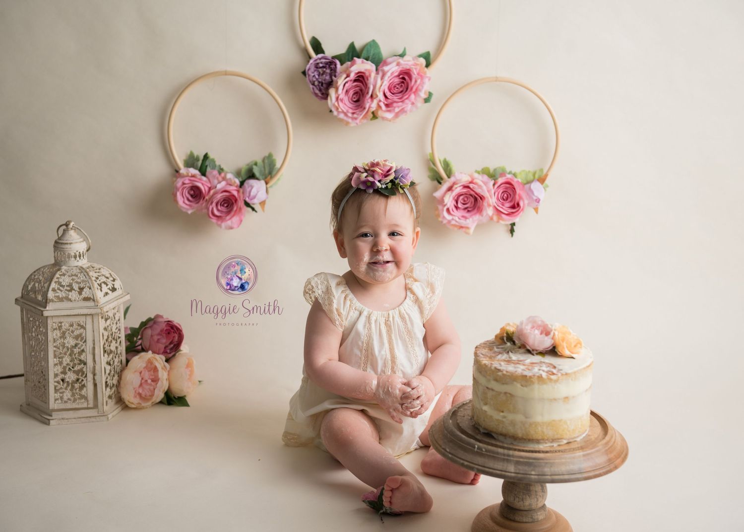 Sweet birthday setup with vintage lantern, floral hoops, rustic cake on wooden stand against neutral backdrop. Cake smash photography
