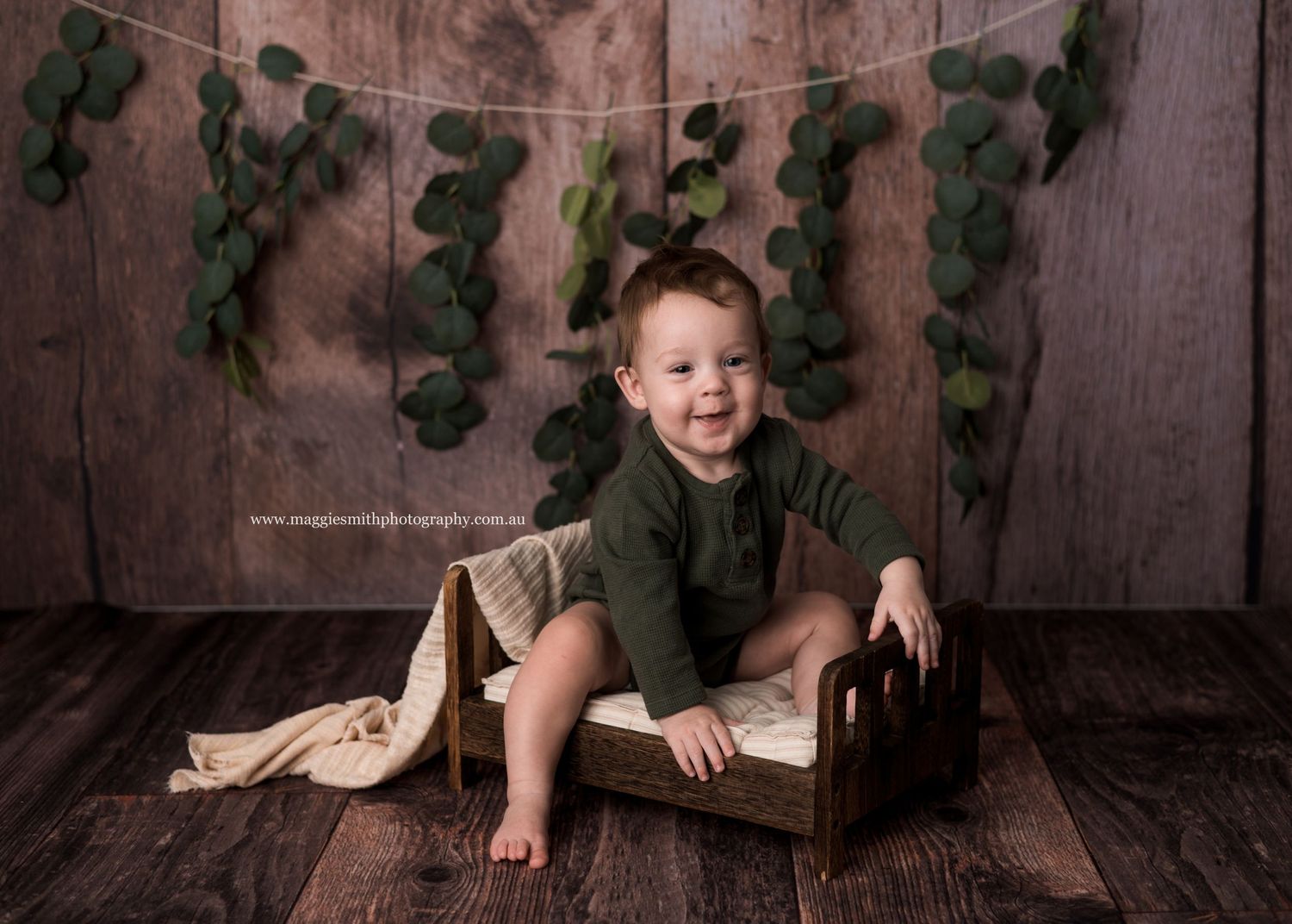 A child sits on a brown wooden prop chair against a rustic backdrop with green foliage decor.
