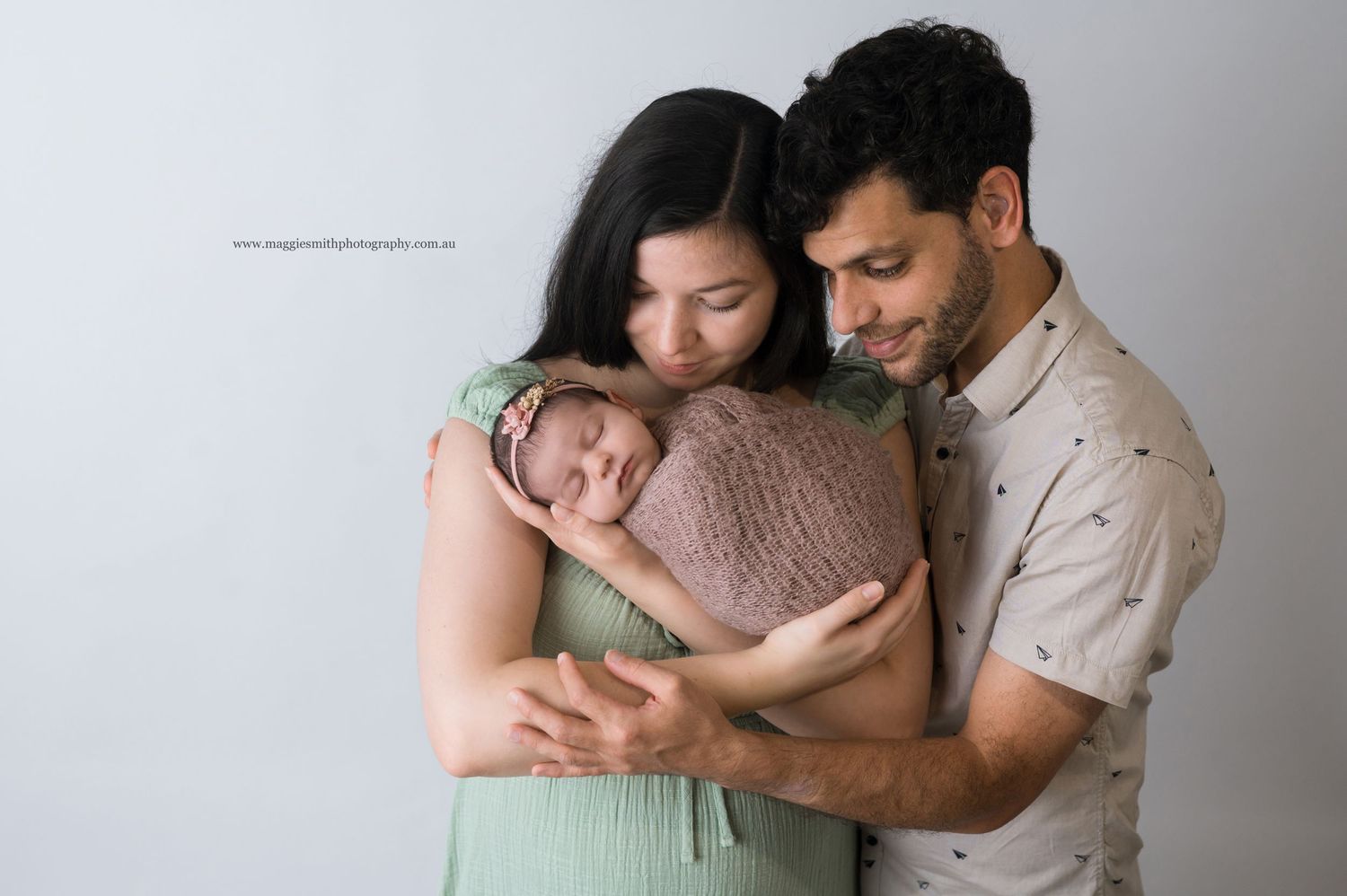 A couple holds their sleeping newborn baby wrapped in a soft brown swaddle against a light gray background.