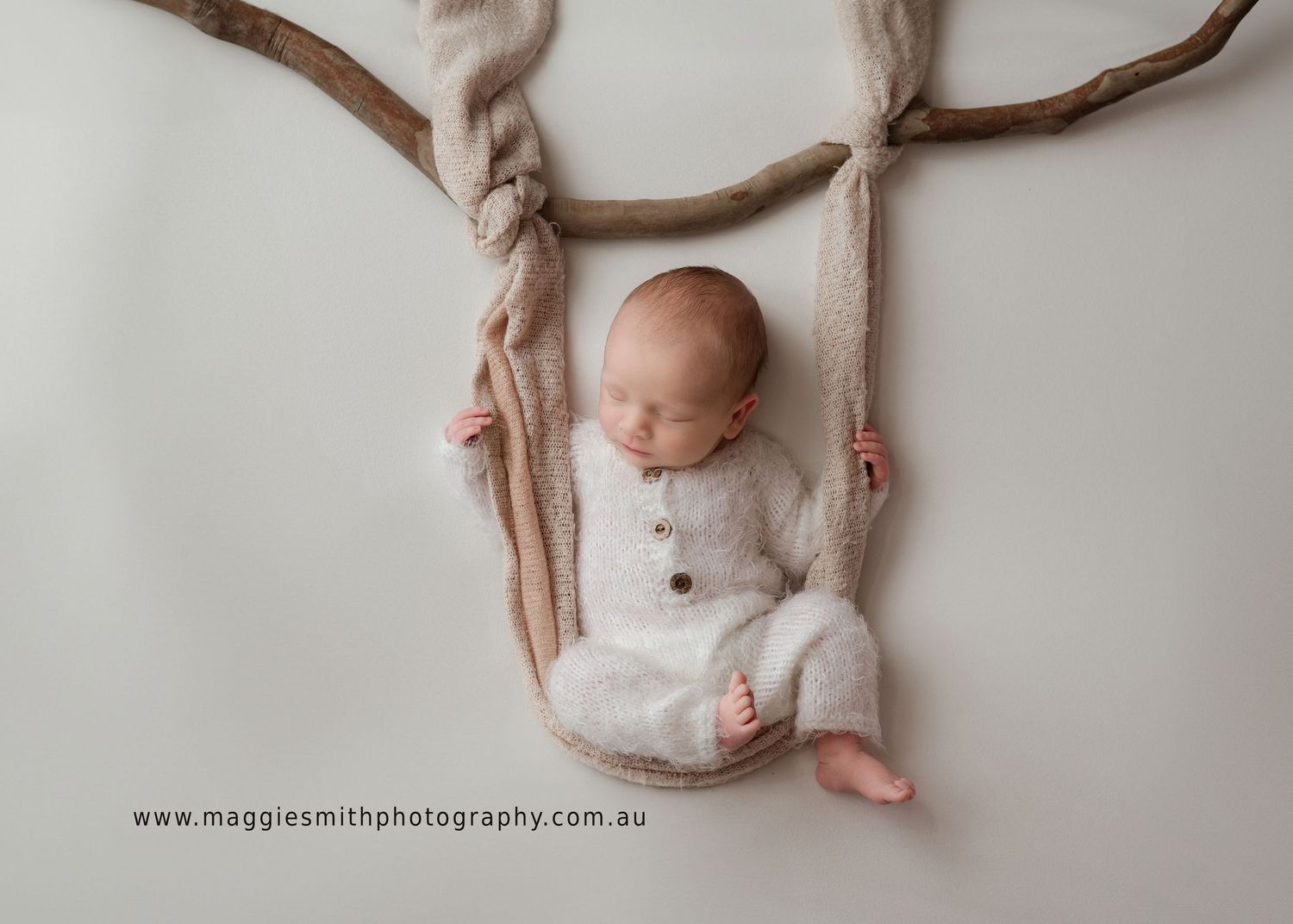 Sleeping newborn in white onesie rests peacefully on fabric swing suspended from brown branch in studio setting.