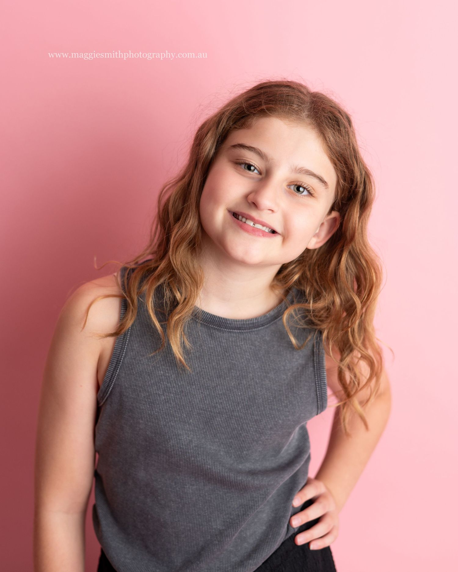 Young person in gray tank top smiling against pink background with wavy brown hair and cheerful expression. Headshot photography