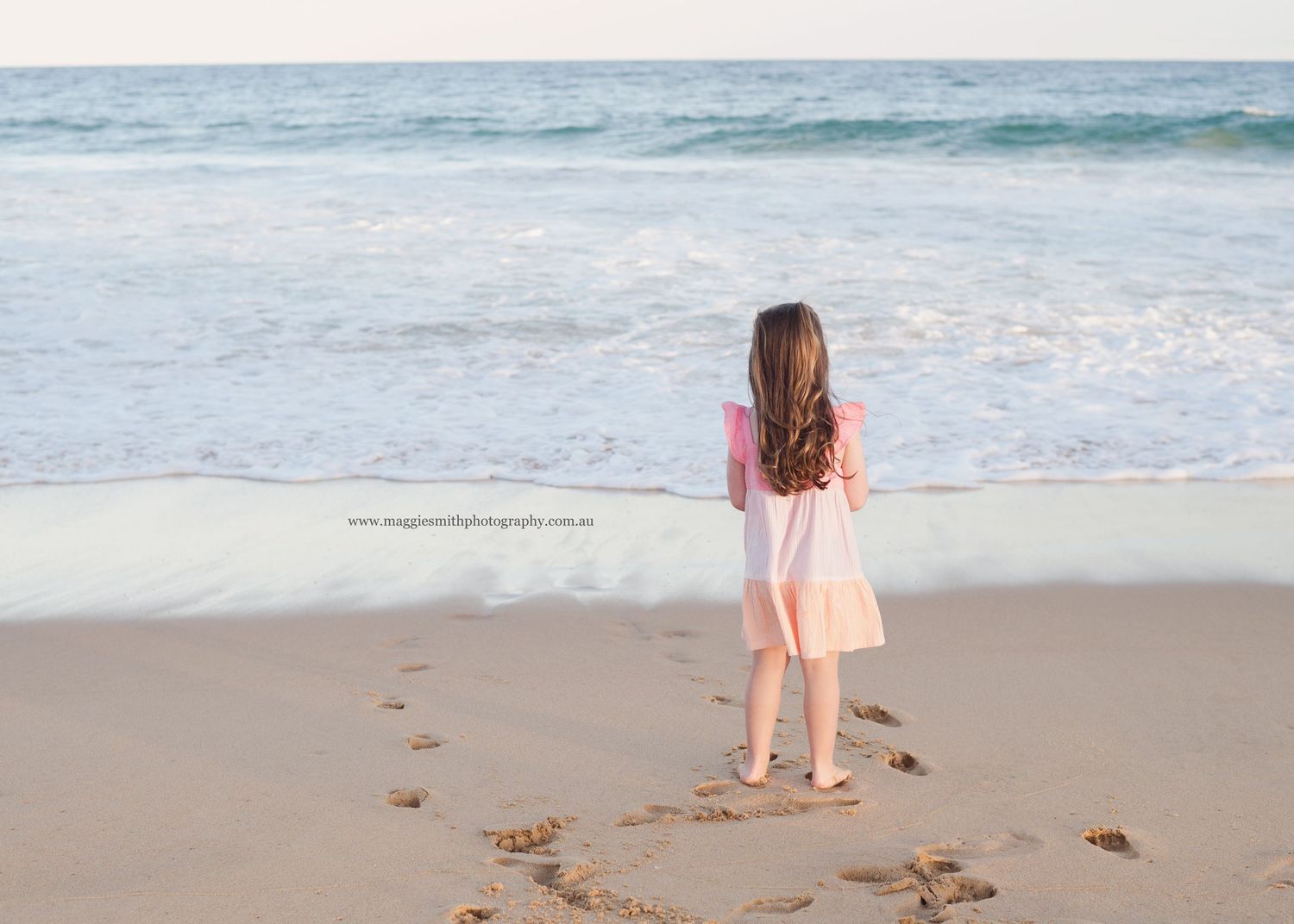 A girl in a pink dress walks along a sandy beach leaving footprints near the ocean waves.