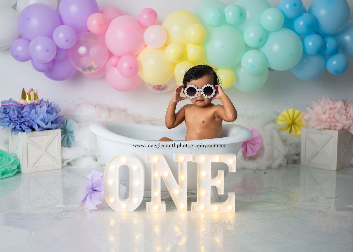 First birthday party setup with illuminated ONE sign and pastel rainbow balloon arch against white backdrop. Cake smash photography