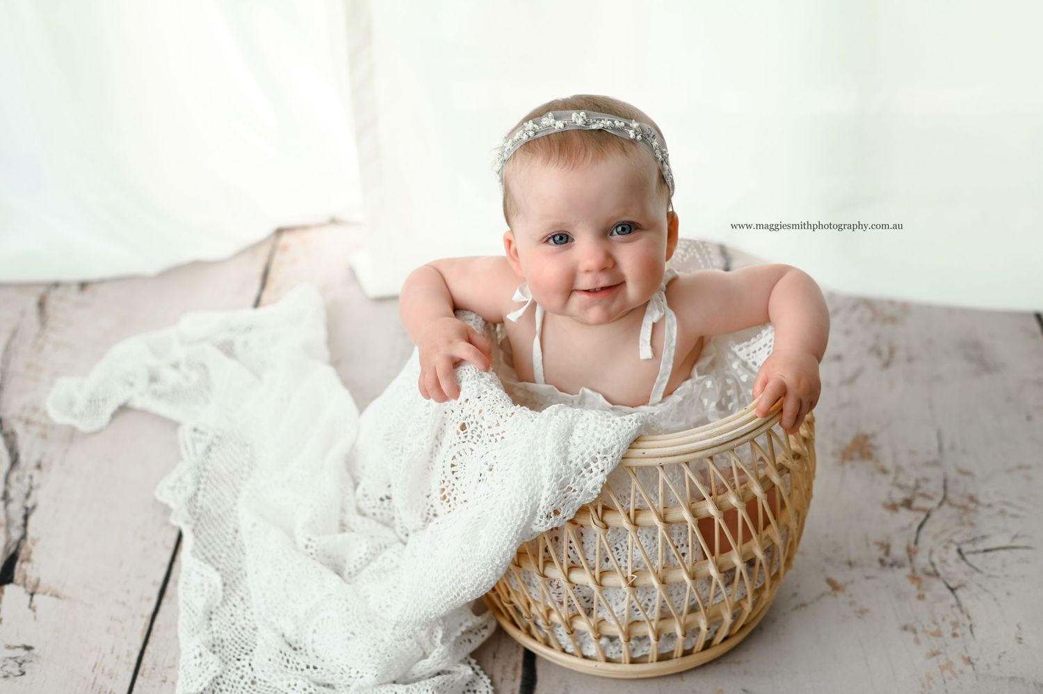 Baby in white dress sits inside woven wicker basket on wooden floor with soft filtered lighting.