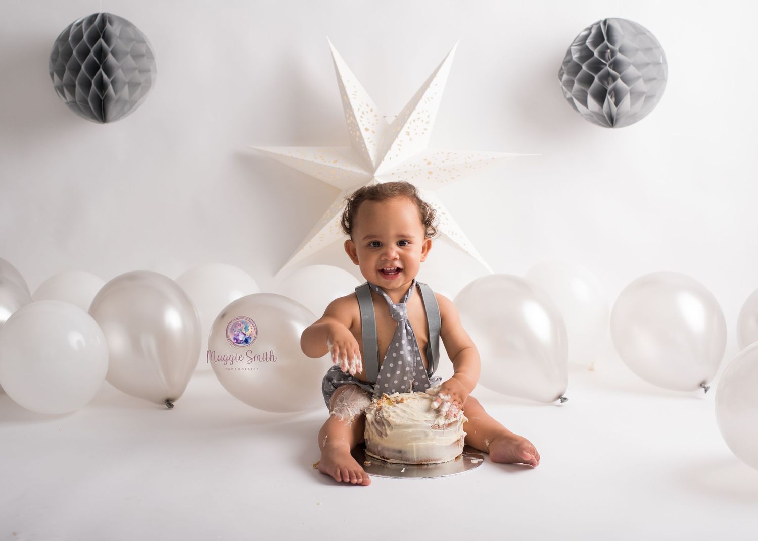 Baby sits on white floor with cake surrounded by white balloons and silver decorative honeycomb balls. Cake smash photography