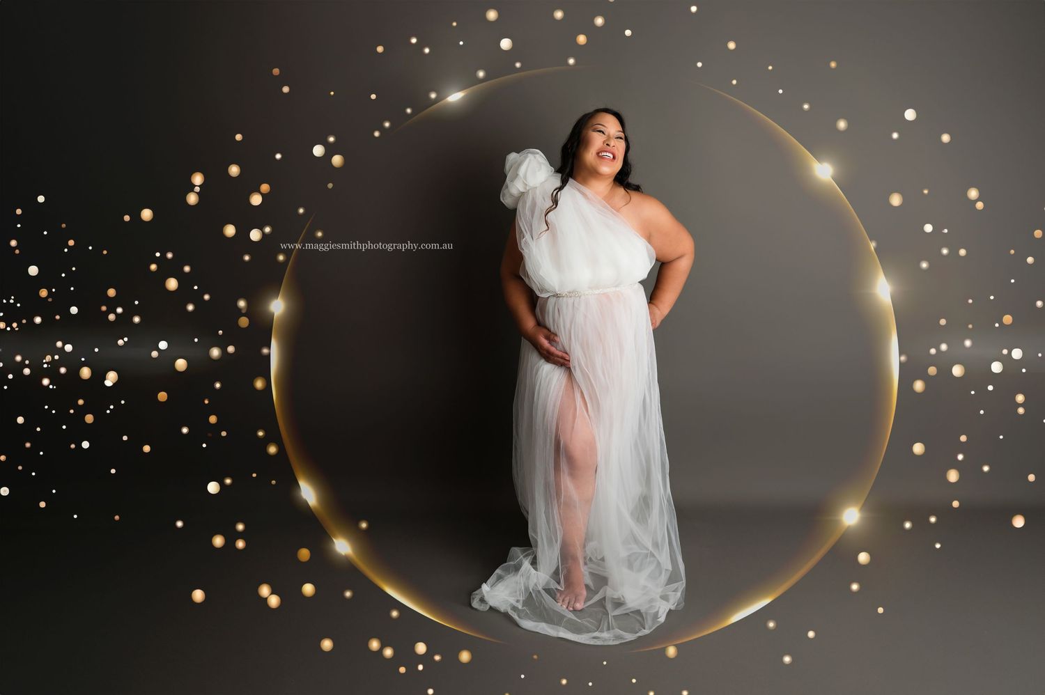 Ethereal maternity portrait with flowing white gown against grey backdrop surrounded by golden sparkles and circular halo.