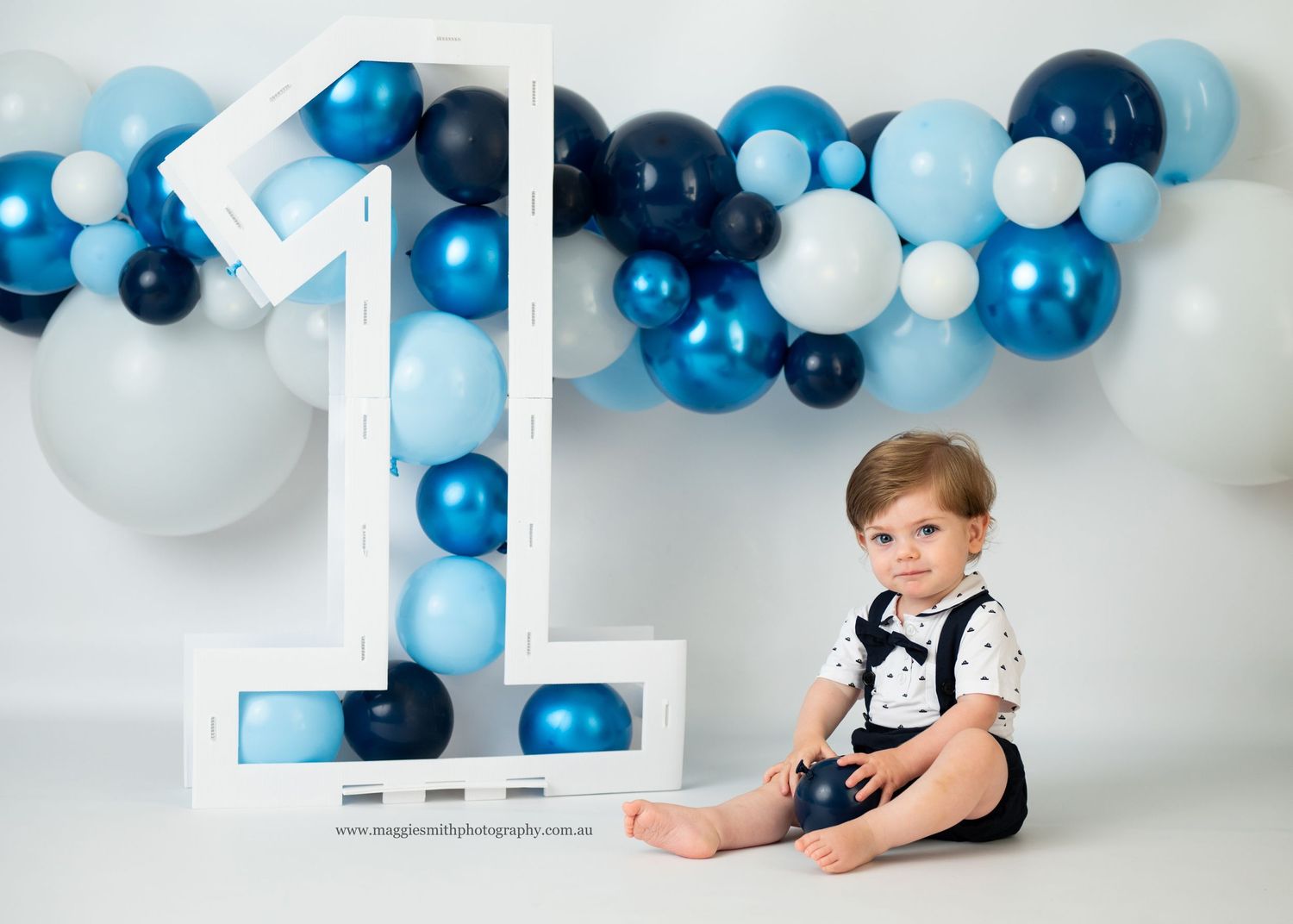 First birthday celebration setup with large white number one and blue balloon arch garland decorations against light background.
