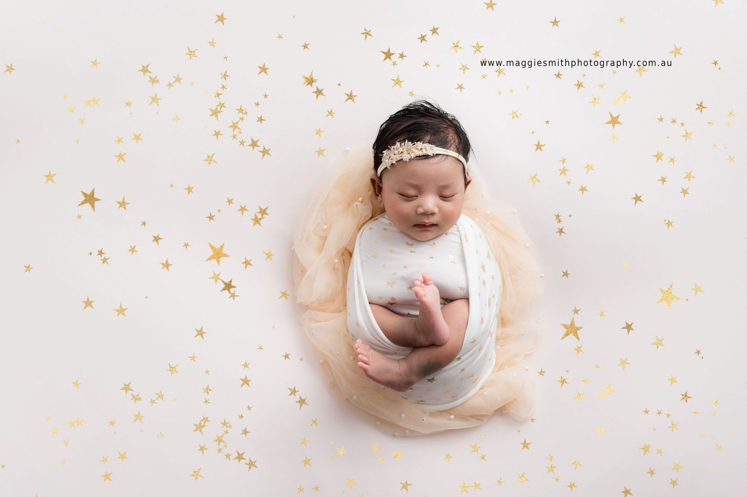 Sleeping newborn wrapped in white fabric lying on cream background with scattered gold star decorations.