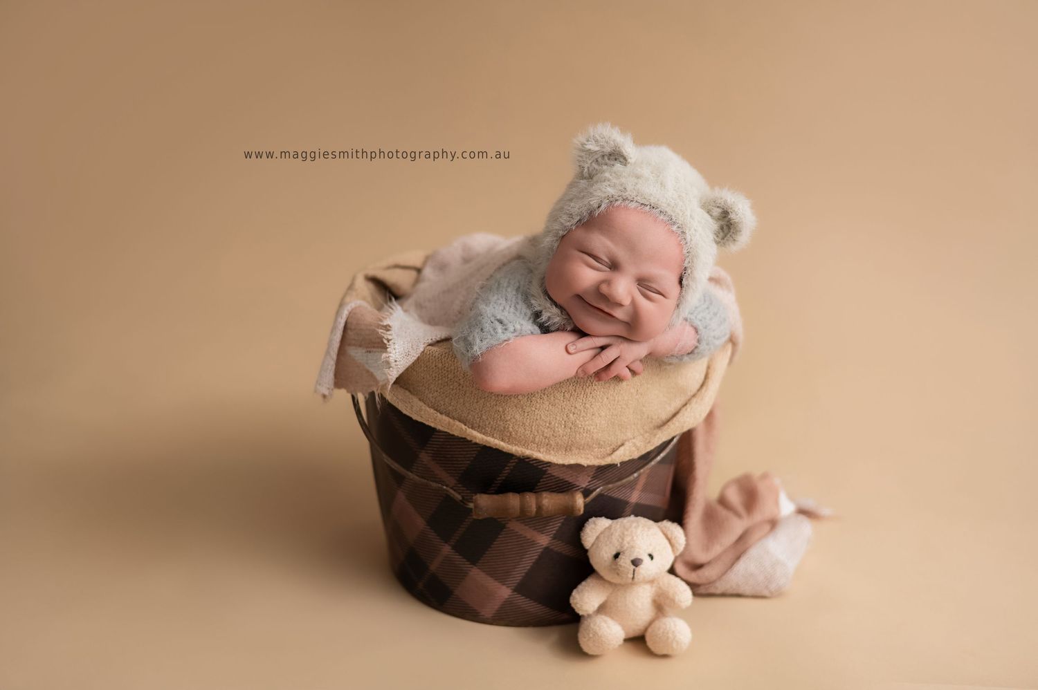 Teddy bear and knit hat arranged in rustic wooden bowl against neutral beige background for newborn photography.