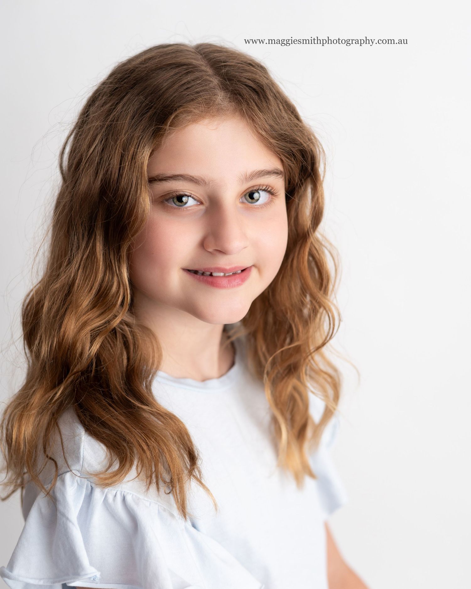 Portrait of a child with wavy light brown hair wearing a white top against a light gray studio background. Headshot photography