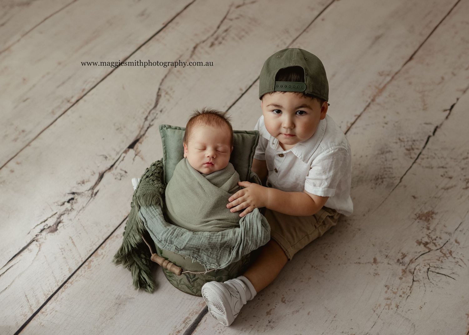 Big brother sitting on wooden floor, with newborn brother swaddled in soft green wrap posed in a bucket during newborn shoot.