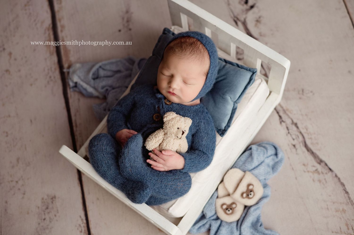 Newborn baby sleeping peacefully in blue outfit while holding small teddy bear in white wooden crate. Newborn photography Central Coast
