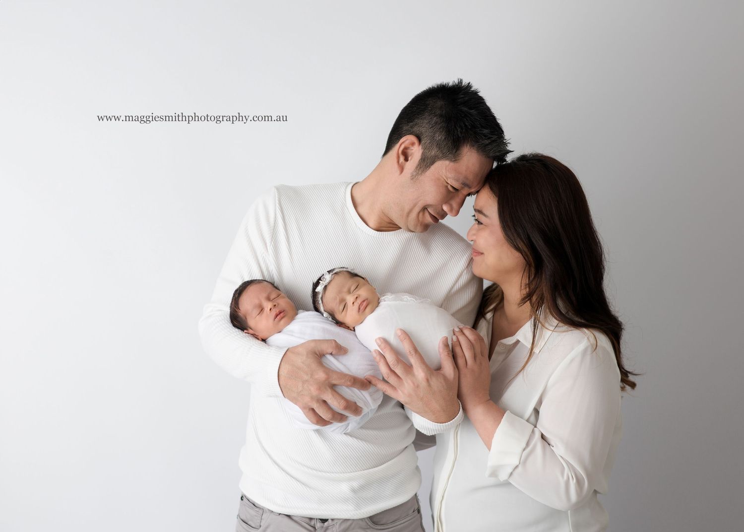 Parents in white tops share a tender moment with their newborn twins in a bright studio setting. Central Coast newborn photography