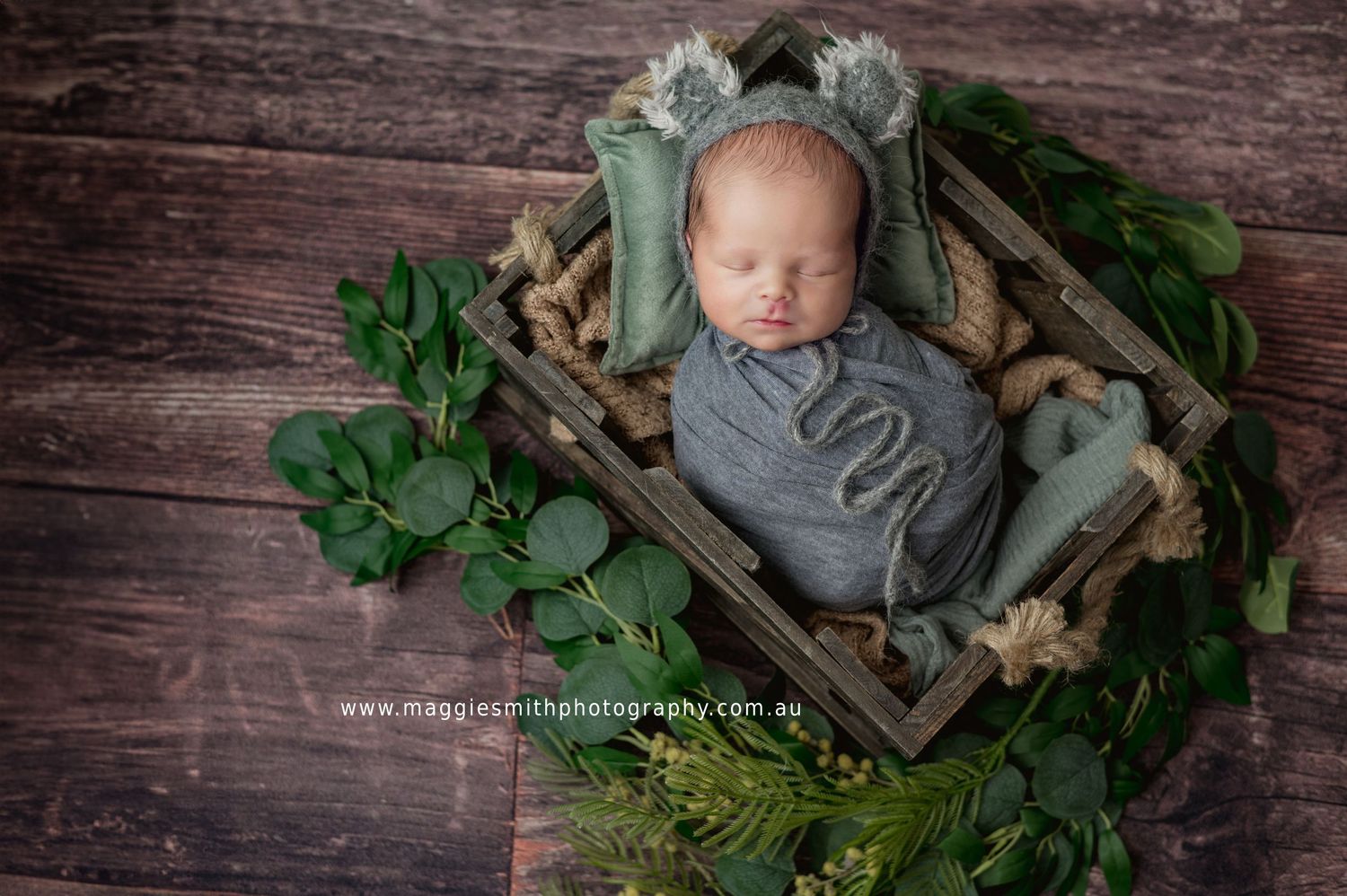 Peaceful sleeping newborn swaddled in gray blanket resting in rustic wooden basket surrounded by green foliage.