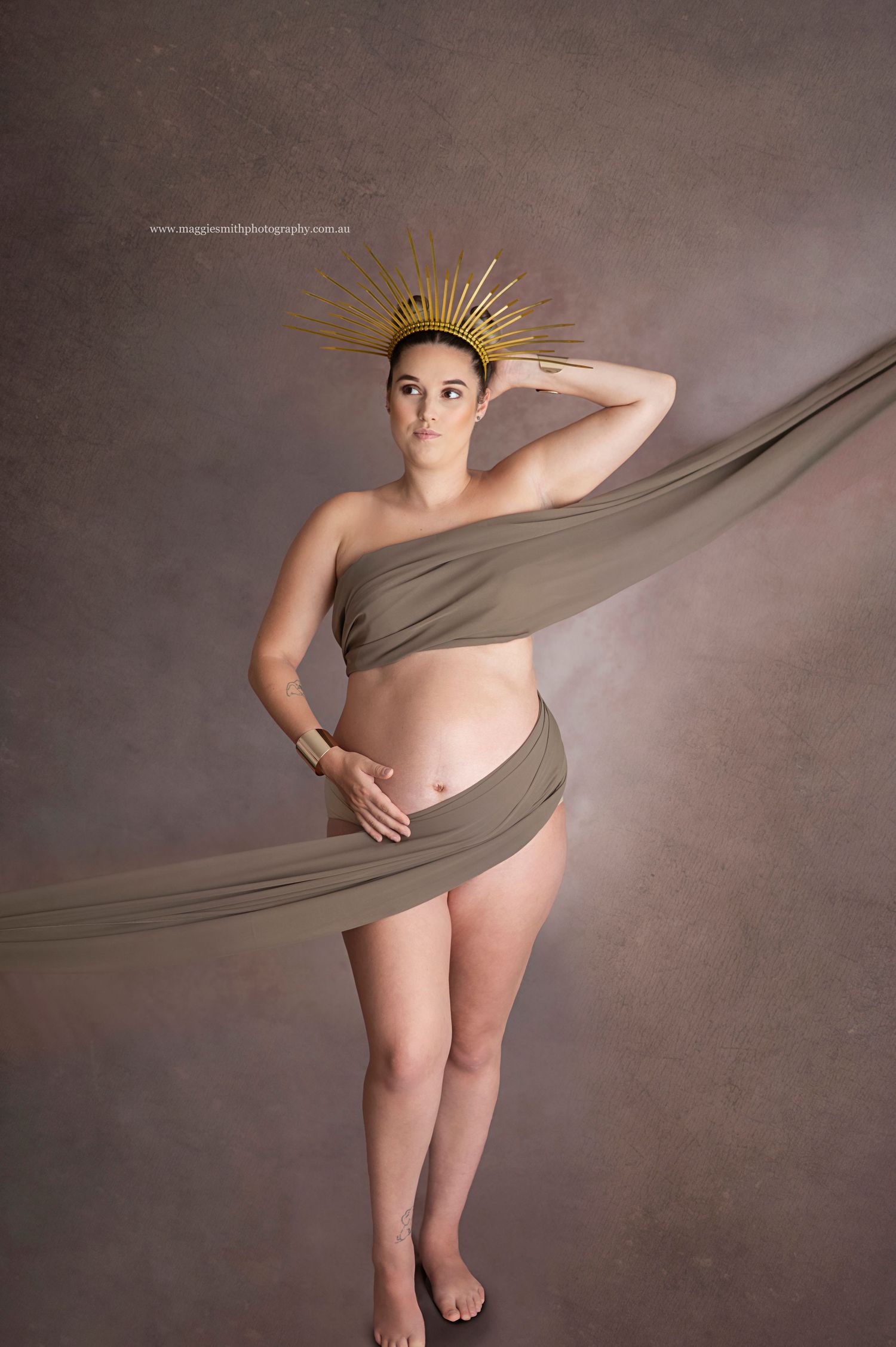 Artistic maternity photo with flowing brown fabric and gold sunburst crown against neutral background.