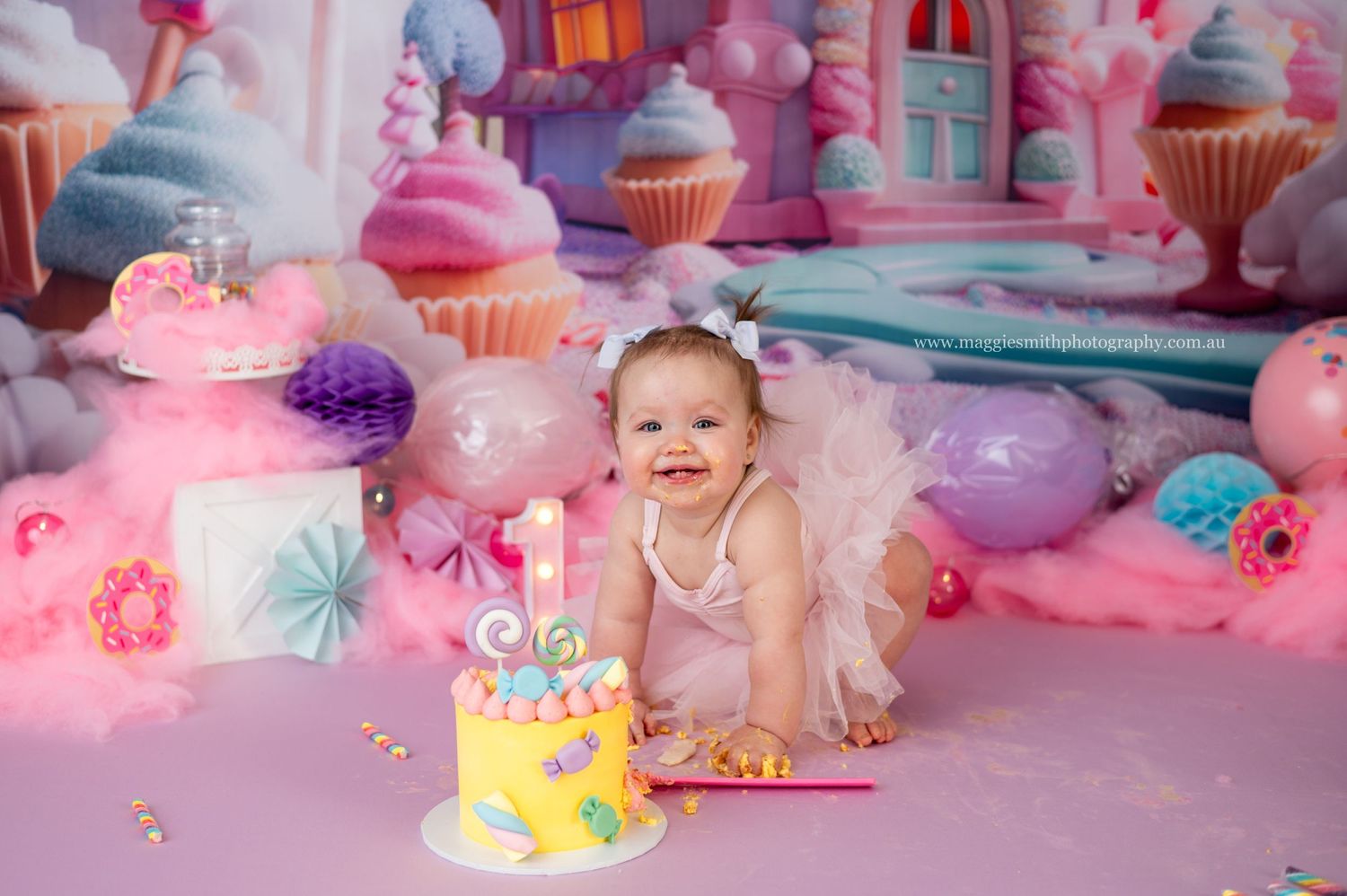 Sweet cake smash photo scene with pastel pink decorations, balloons and cupcakes surrounding a yellow birthday cake. Candyland theme