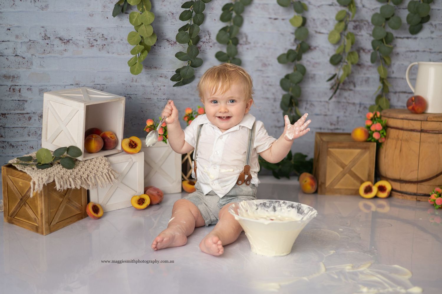 A joyful child sits on a floor with wooden crates, peaches and greenery in a rustic styled photography setup.