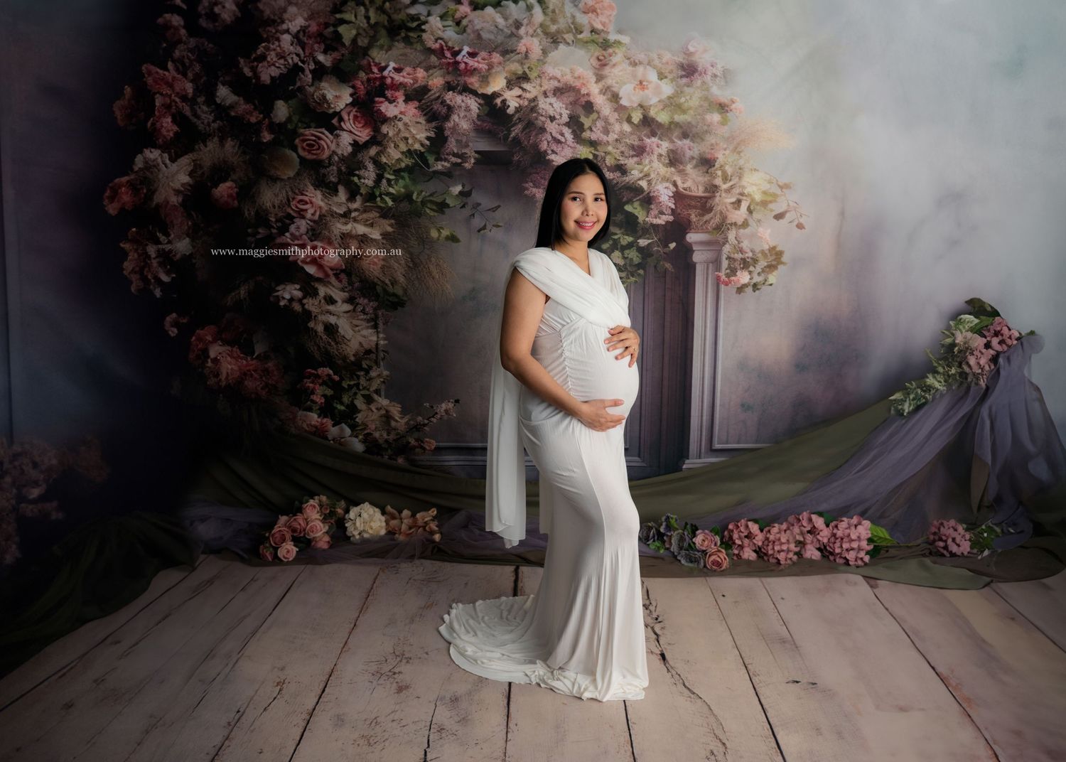 Maternity photoshoot in white gown against dreamy pink floral backdrop with soft lighting and wooden floor.