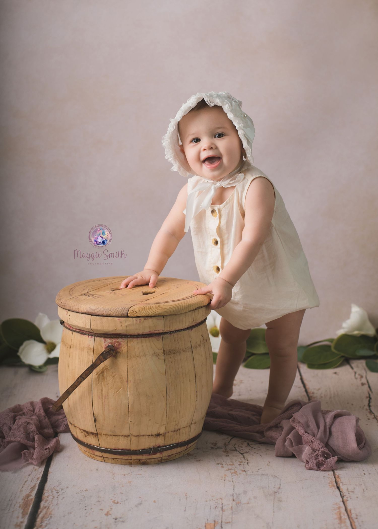 Young child in white dress and bonnet leans on vintage wooden barrel in rustic setting with soft pink backdrop.