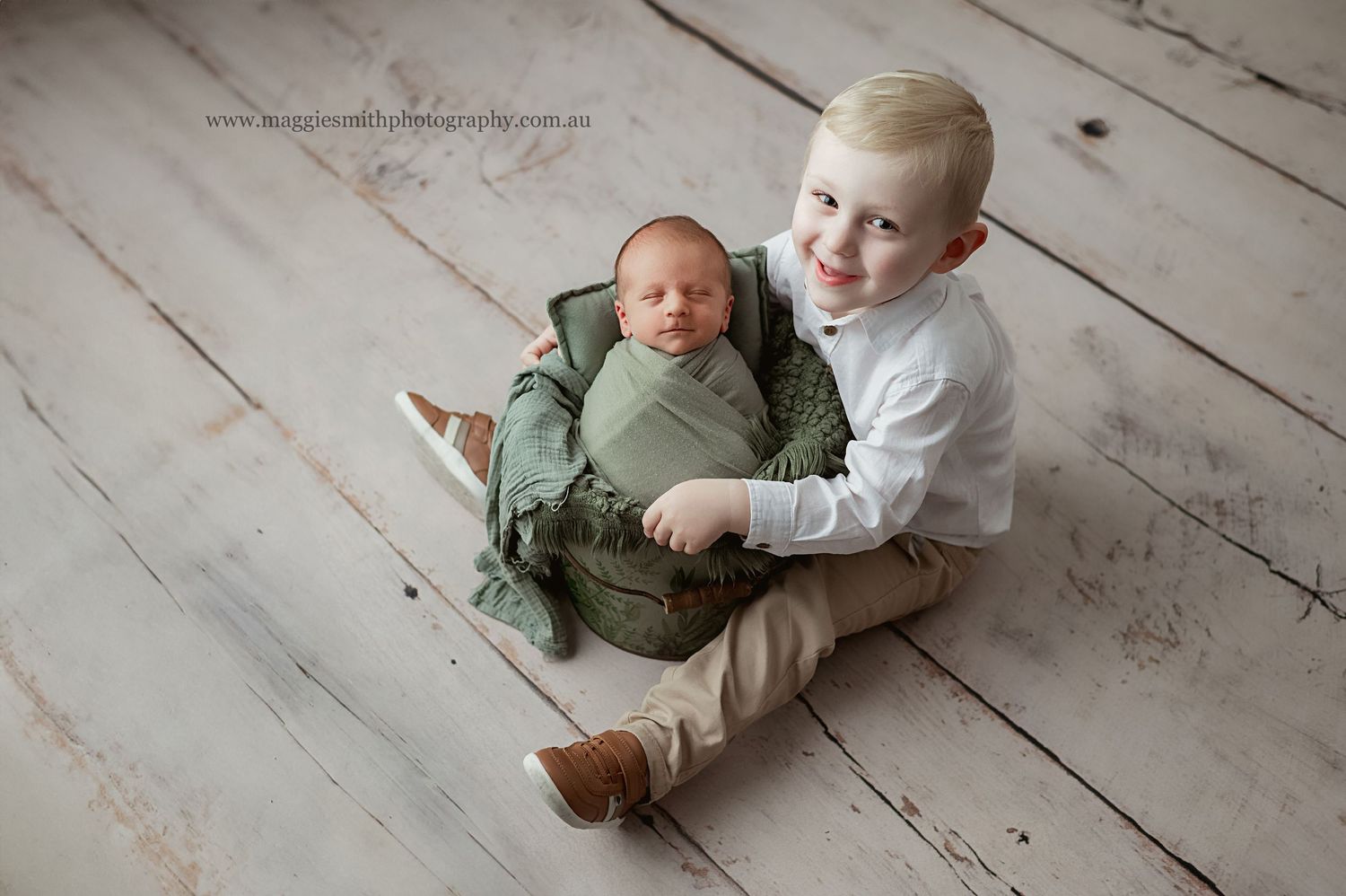 Young child sits on rustic wood floor with swaddled newborn baby in soft green wrap. Newborn photography Central Coast