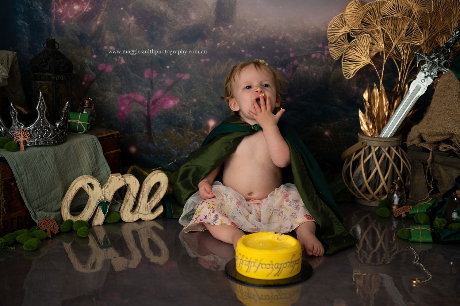 A cake smash photo shoot with gold 'one' lettering, decorative elements, and a ring cake on a dark backdrop. Lord of the Ring theme.