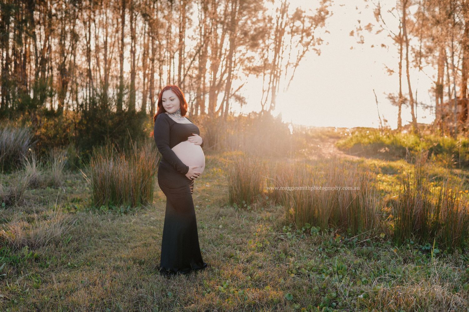 A pregnant silhouette stands in a long black dress against golden sunlight filtering through trees in a serene outdoor setting. Maternity.
