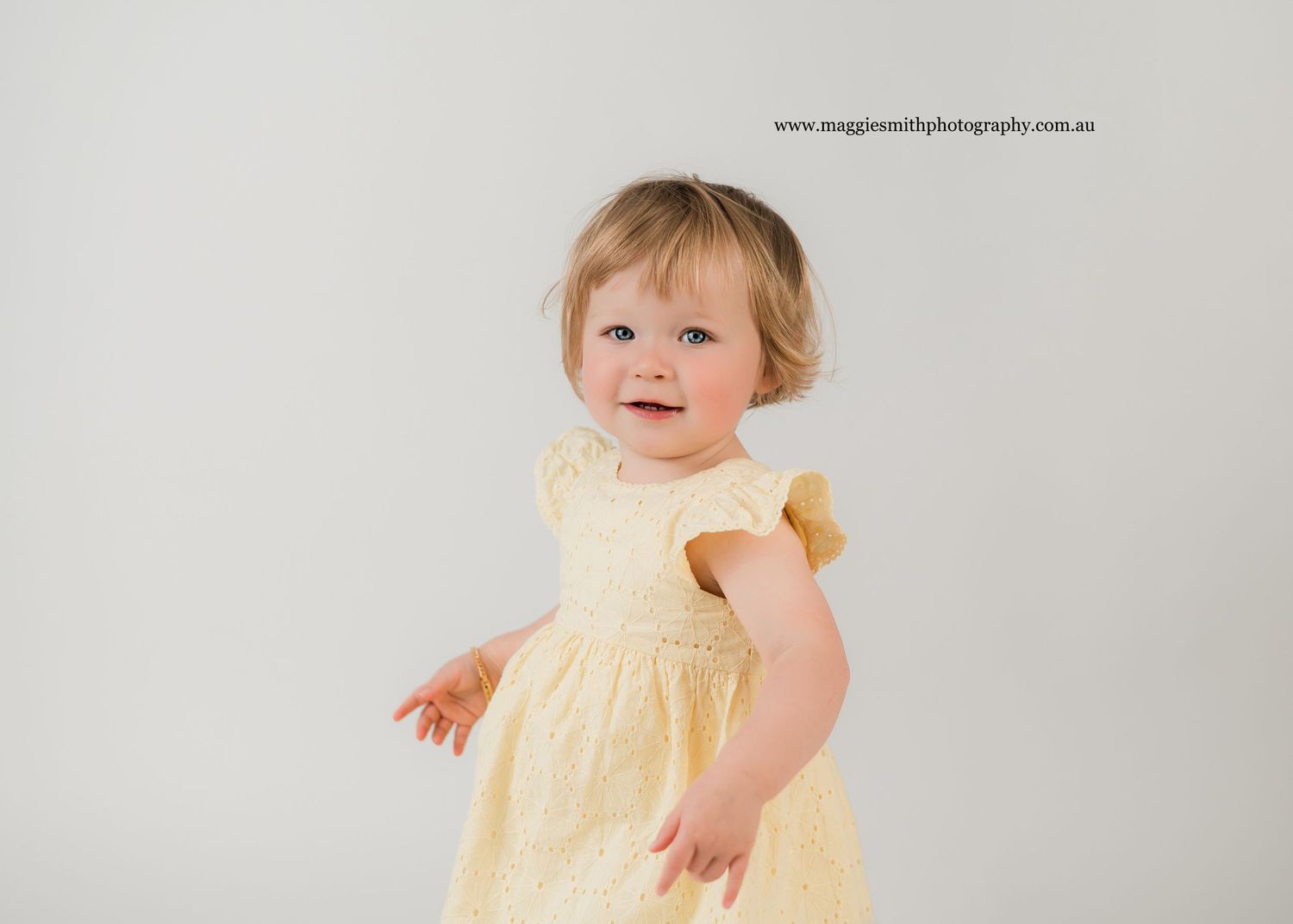 Baby wearing a pale yellow dress smiles while sitting against a white backdrop in a professional studio setting. Kids photography
