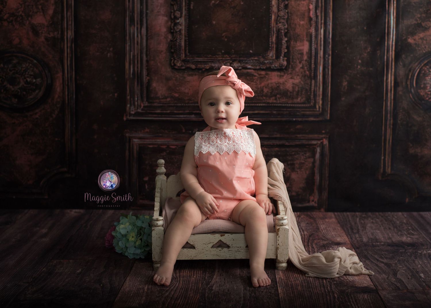 A baby in a peach romper sits on a small wooden bench against a dark vintage door backdrop.