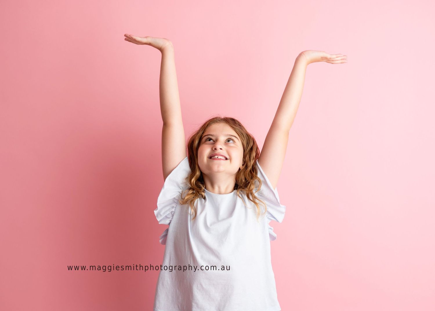 Child in white t-shirt jumps with raised arms and joyful expression against pink background. Headshot photography
