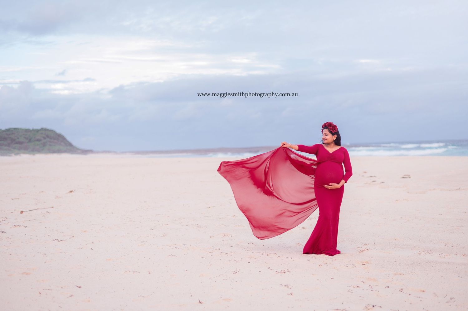Pregnant woman with a flowing red dress stands gracefully on a beach with rolling waves and cloudy skies in the background.