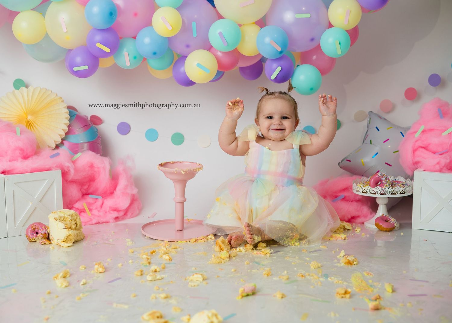 Colorful birthday party setup with pastel balloons, tulle dress, and gold confetti scattered on white floor. Cake smash photography