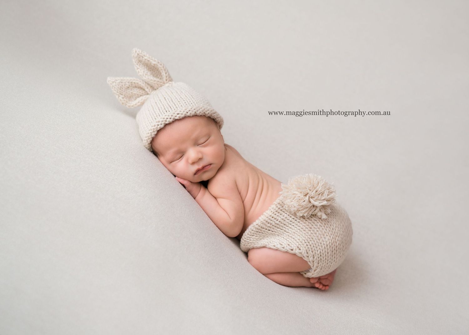 Sleeping newborn wearing soft knitted bunny hat and pants curled up on white backdrop in sweet pose.