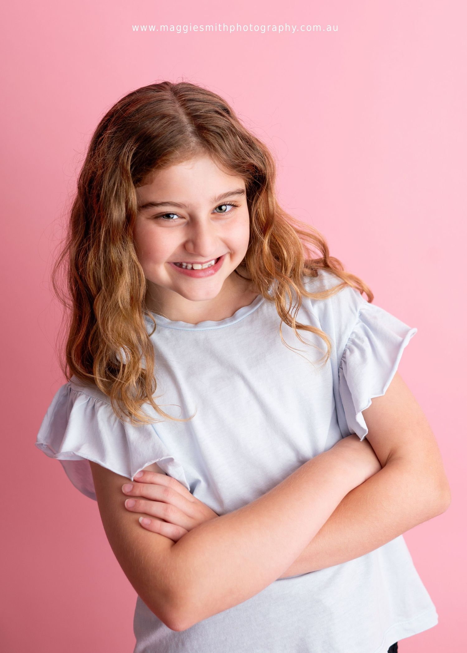 Young child in white t-shirt smiling with arms crossed against pink background. Headshot photography