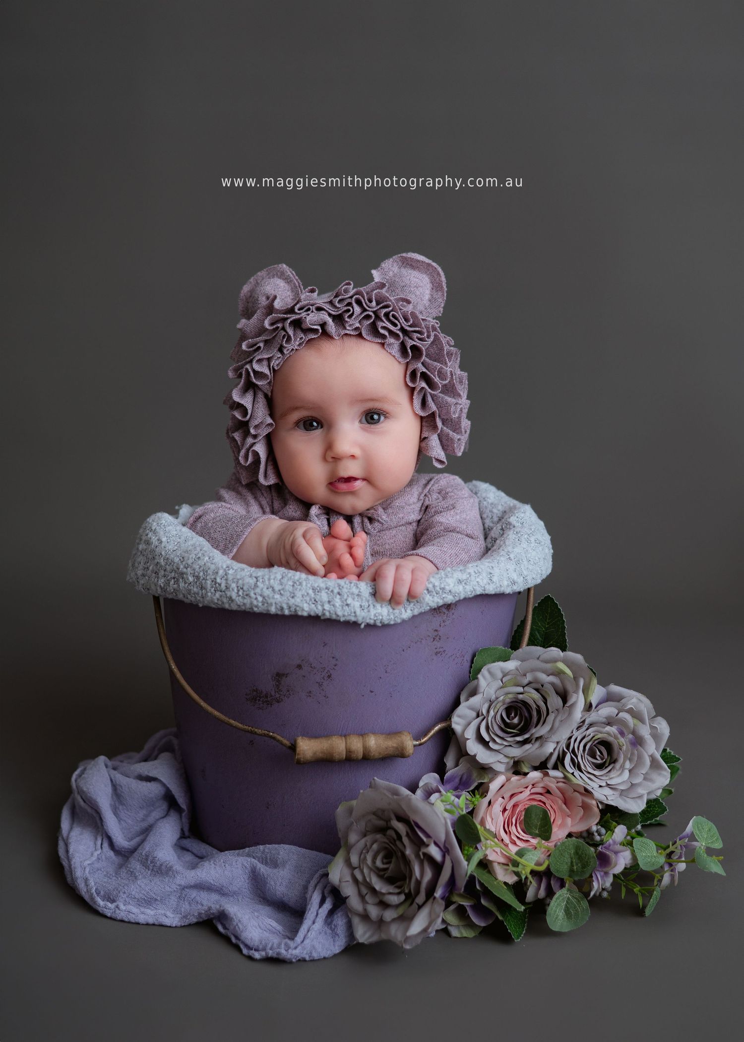 Baby in purple knitted bonnet sits in vintage purple container with gray roses and greenery arrangements beside it.