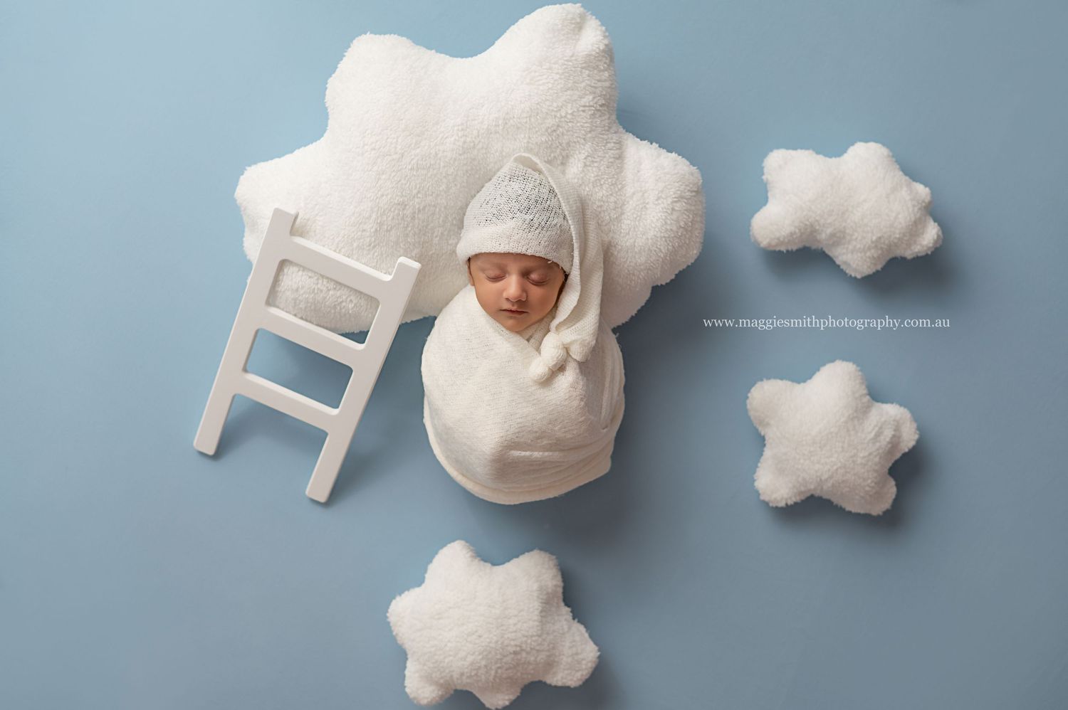 Creative newborn photography with white cloud props and tiny ladder against light blue background.