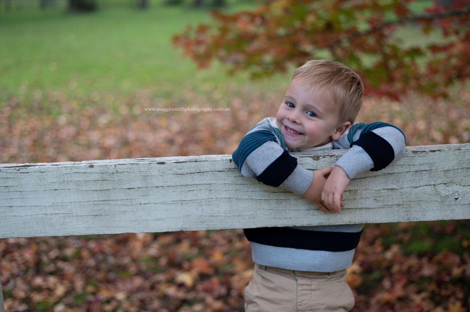 A young child in a striped sweater leans on a white wooden fence rail with autumn leaves in the background.