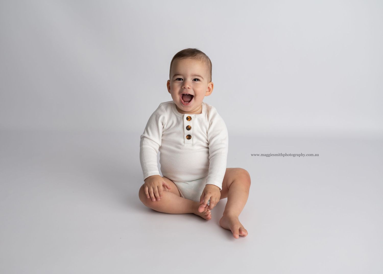Happy baby wearing white henley bodysuit sits on light gray backdrop during professional photo session.