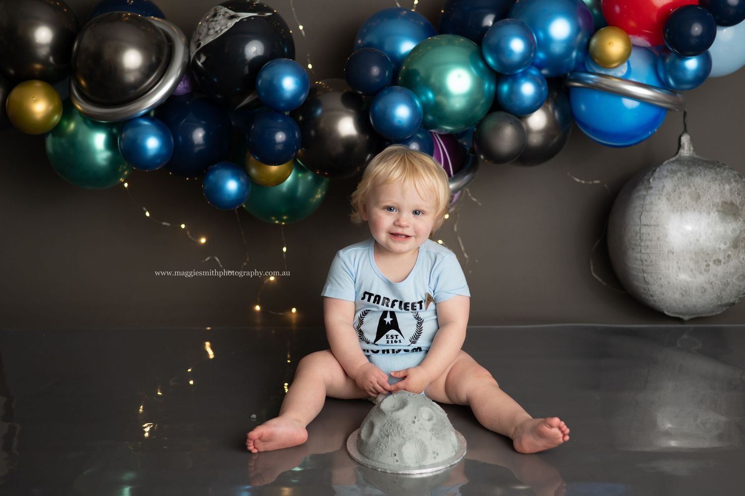 A colorful space-themed cake smash setup with silver planets, metallic balloons, and string lights against a gray backdrop.