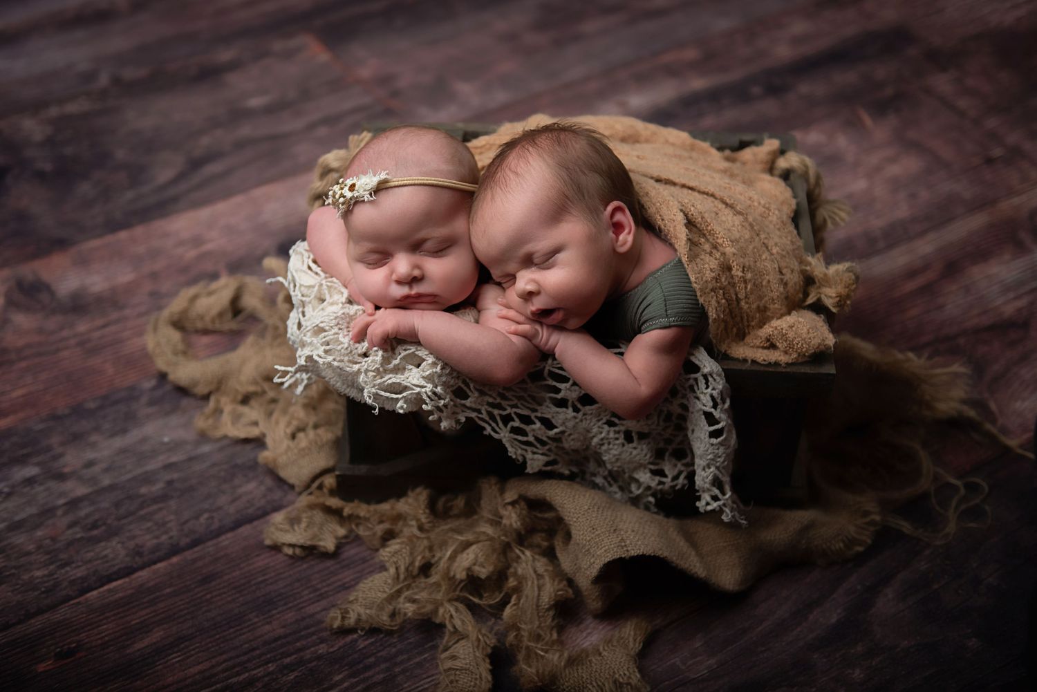 Newborn twins sleeping peacefully on brown lace with delicate floral headpiece and neutral wrapping.