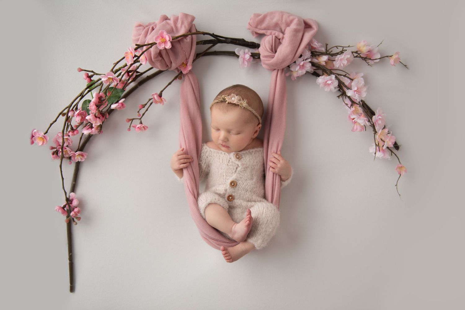 Newborn baby sleeping peacefully in pink fabric swing with cherry blossoms and branches creating an arch above.