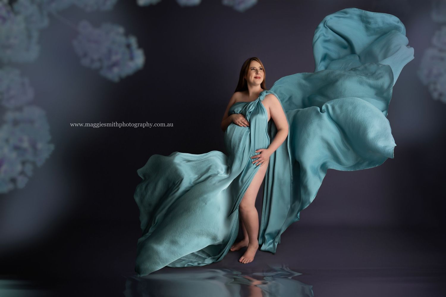 A flowing aqua maternity gown billows dramatically against a dark studio backdrop during a pregnancy photoshoot.