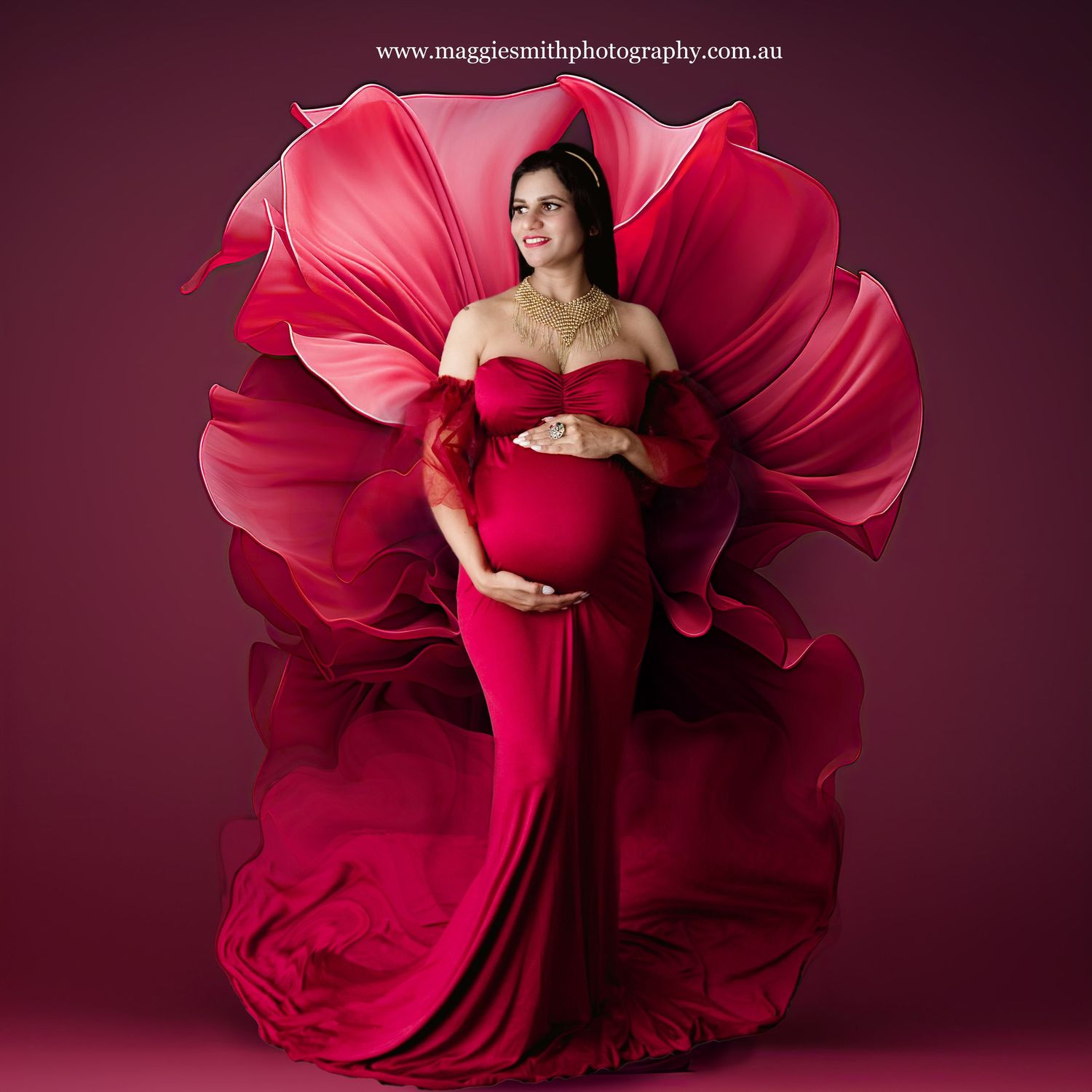 Red maternity photograph featuring an elegant gown with dramatic flowing fabric creating a flower-like backdrop against burgundy.