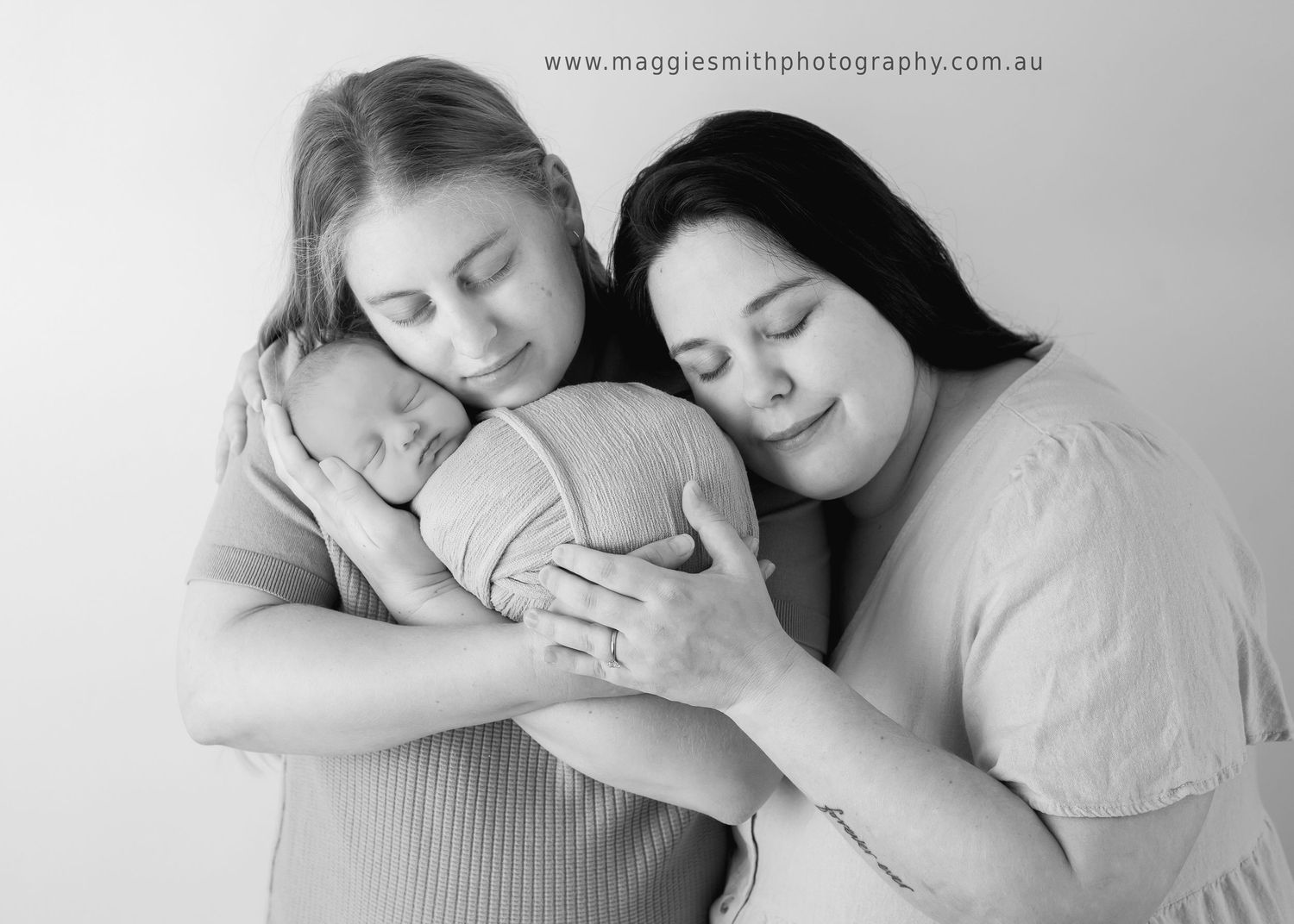 An intimate black and white portrait of an LGBTQ+ couple cradling a sleeping newborn baby with tender affection.