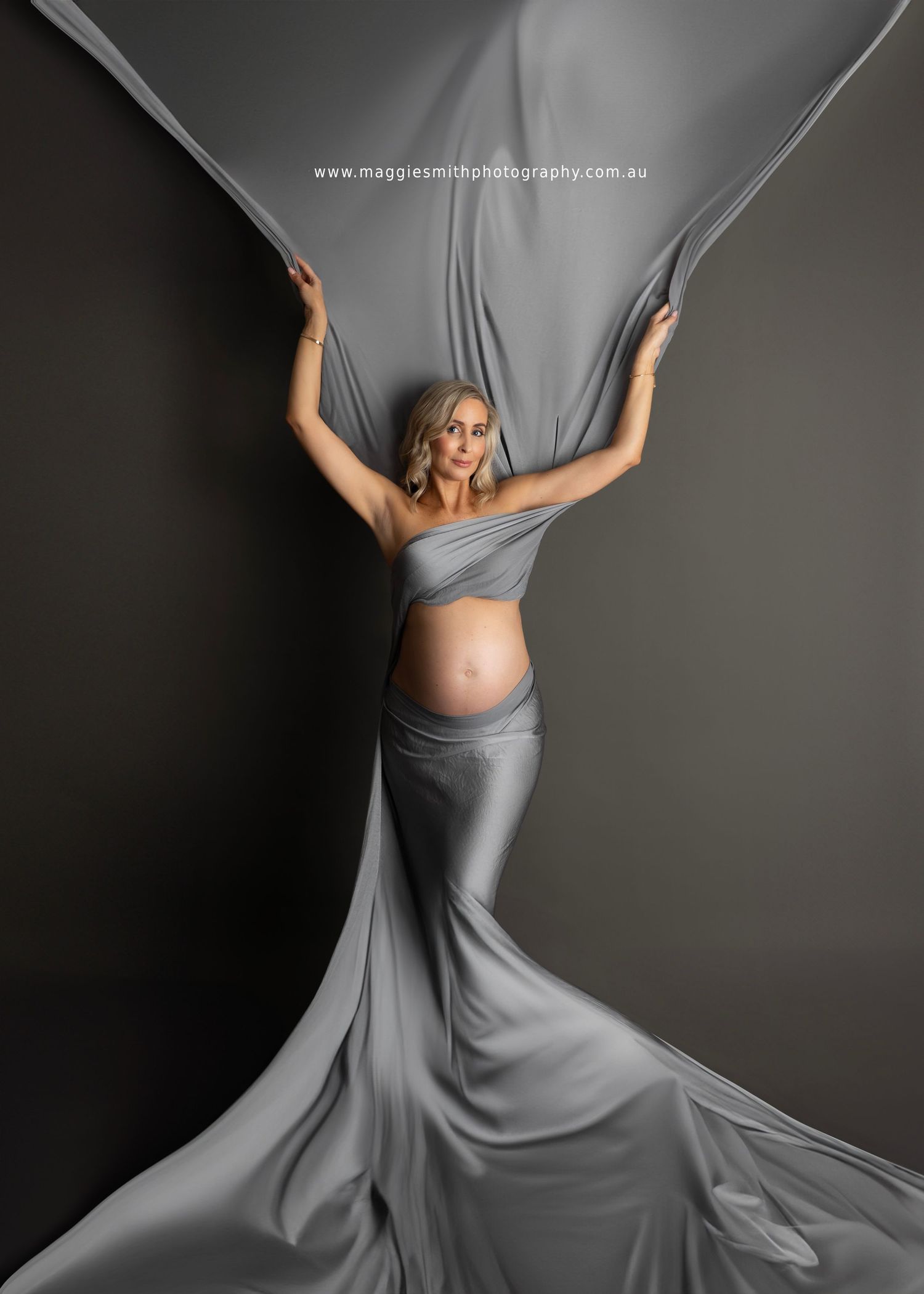 A pregnant woman in a flowing gray dress twirls gracefully against a dark background in an artistic maternity photo series. Maternity photo