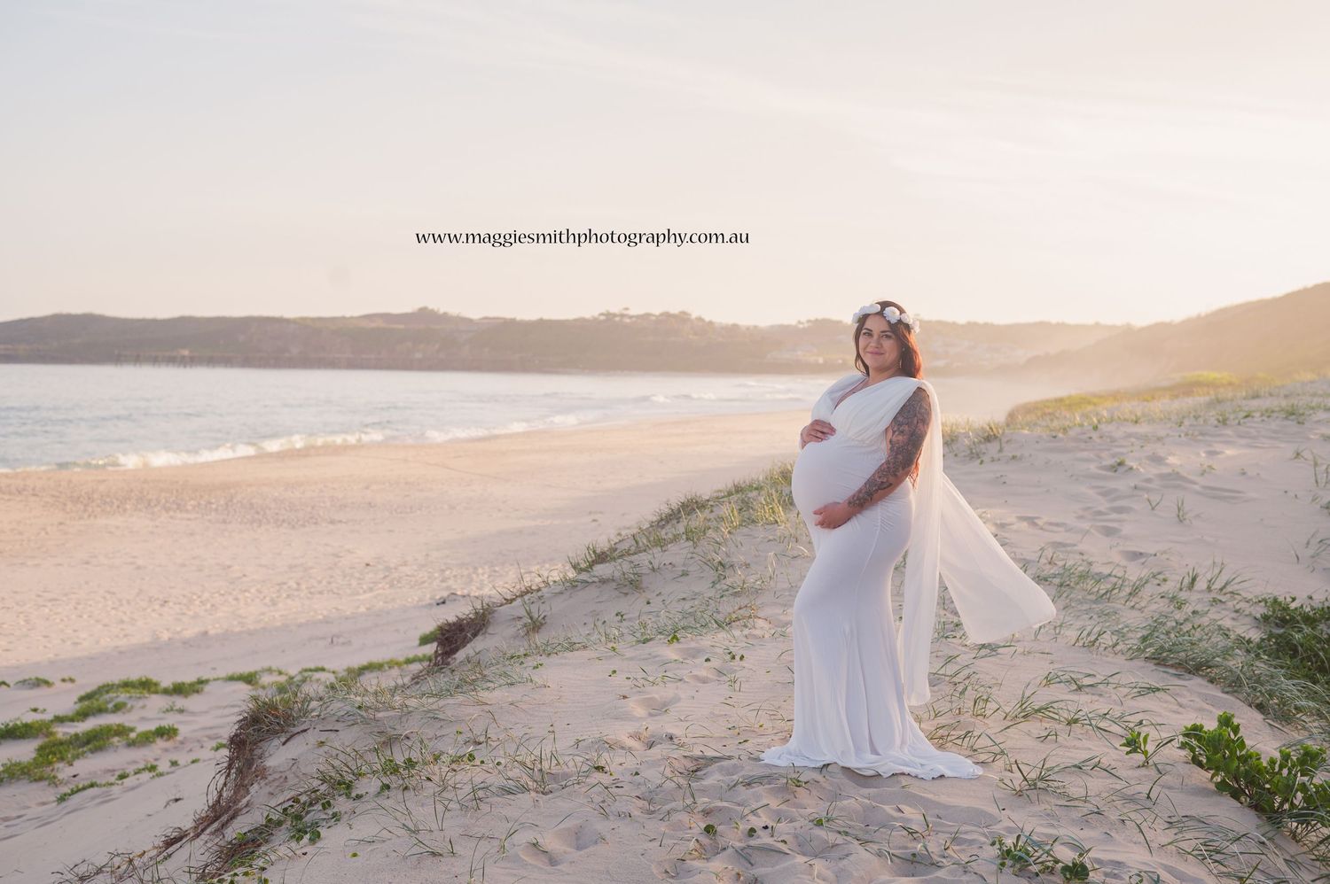 Pregnant lady in flowing white gown stands on sandy dunes overlooking a serene beach at sunset.