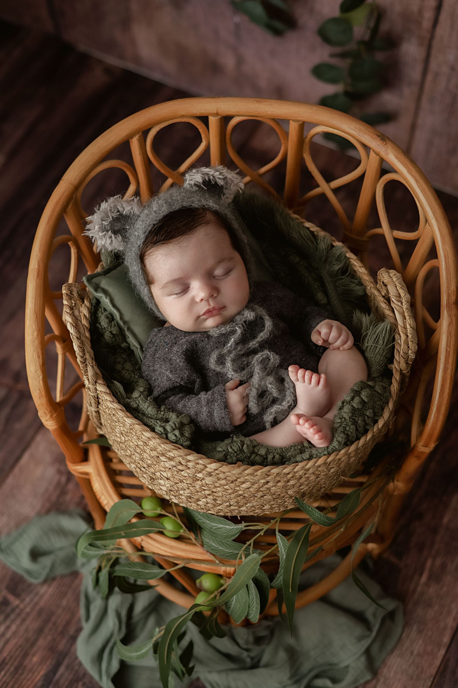 Newborn baby sleeping peacefully in rattan chair with olive green blanket and furry koala bear hat during professional newborn photoshoot.