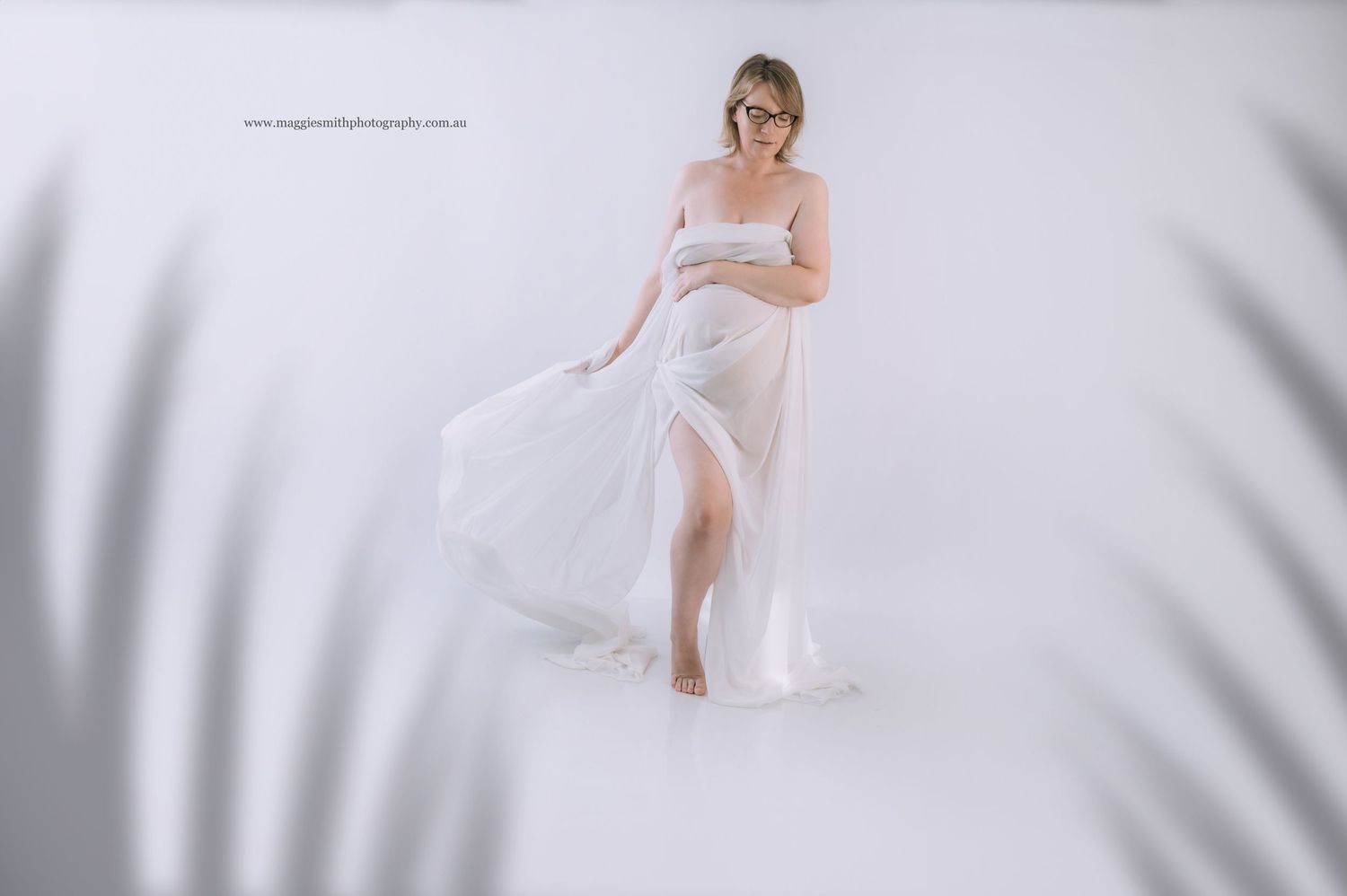 A figure in a flowing white off-shoulder maternity dress poses elegantly against a soft gray background.